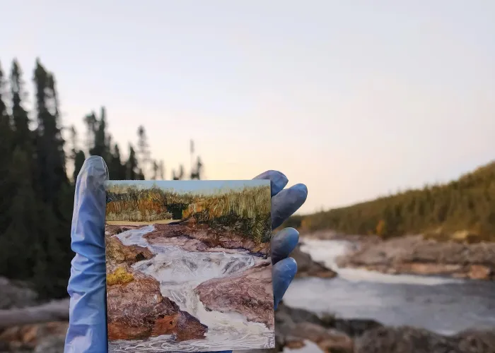 Blue-gloved hand holding small painting of river that is also in the backrgound