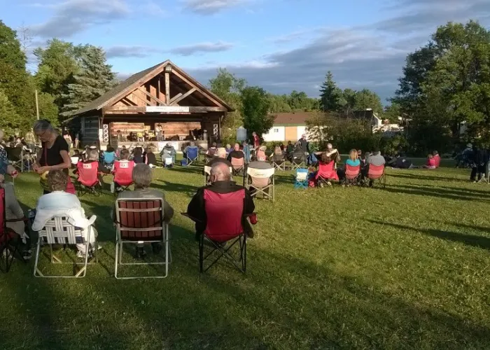 Audience in lawn chairs and sitting on grass watches band play in wooden building stage